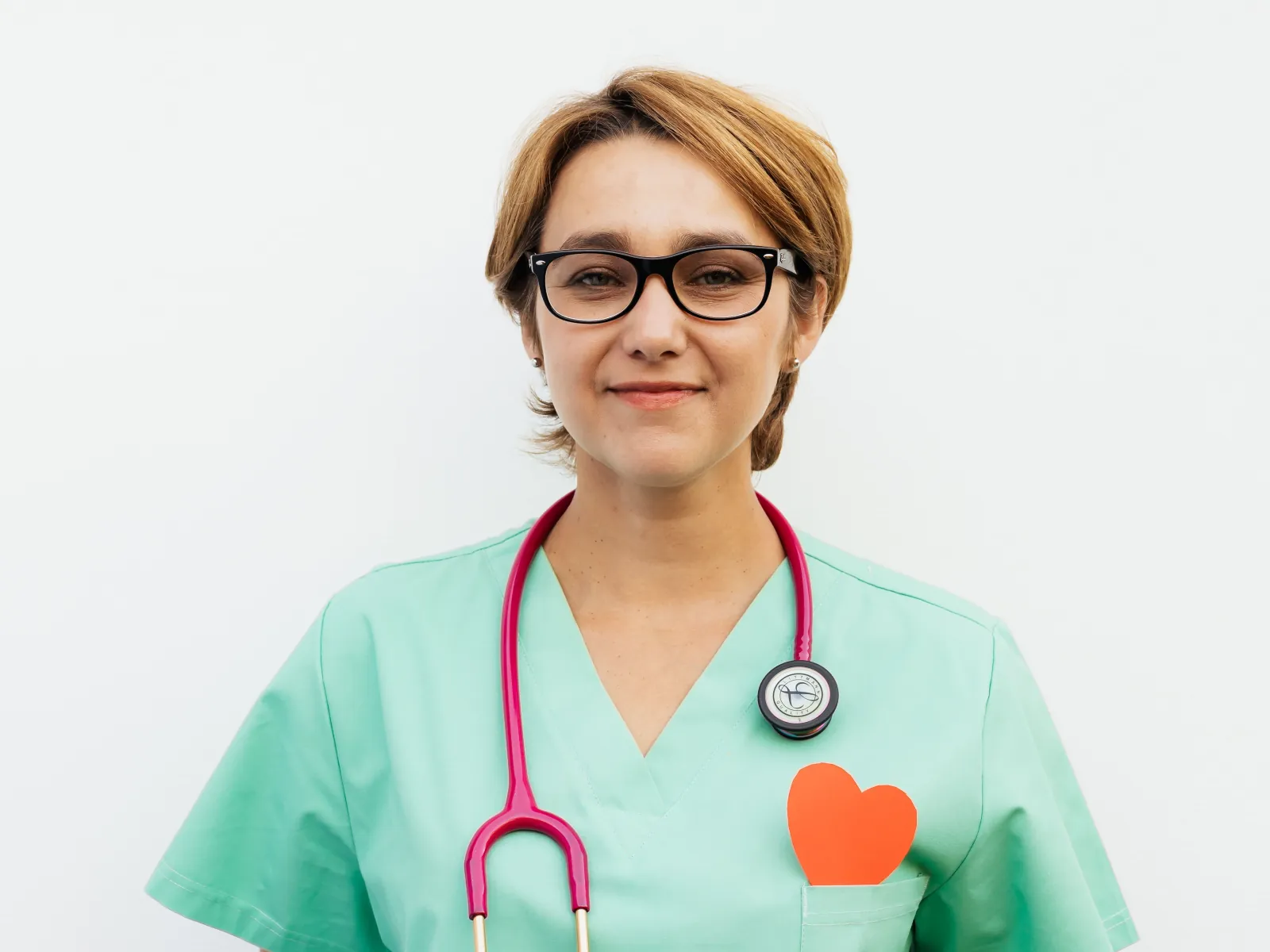 Mujer con gafas y uniforme verde sonriendo con un corazón de papel naranja en el bolsillo